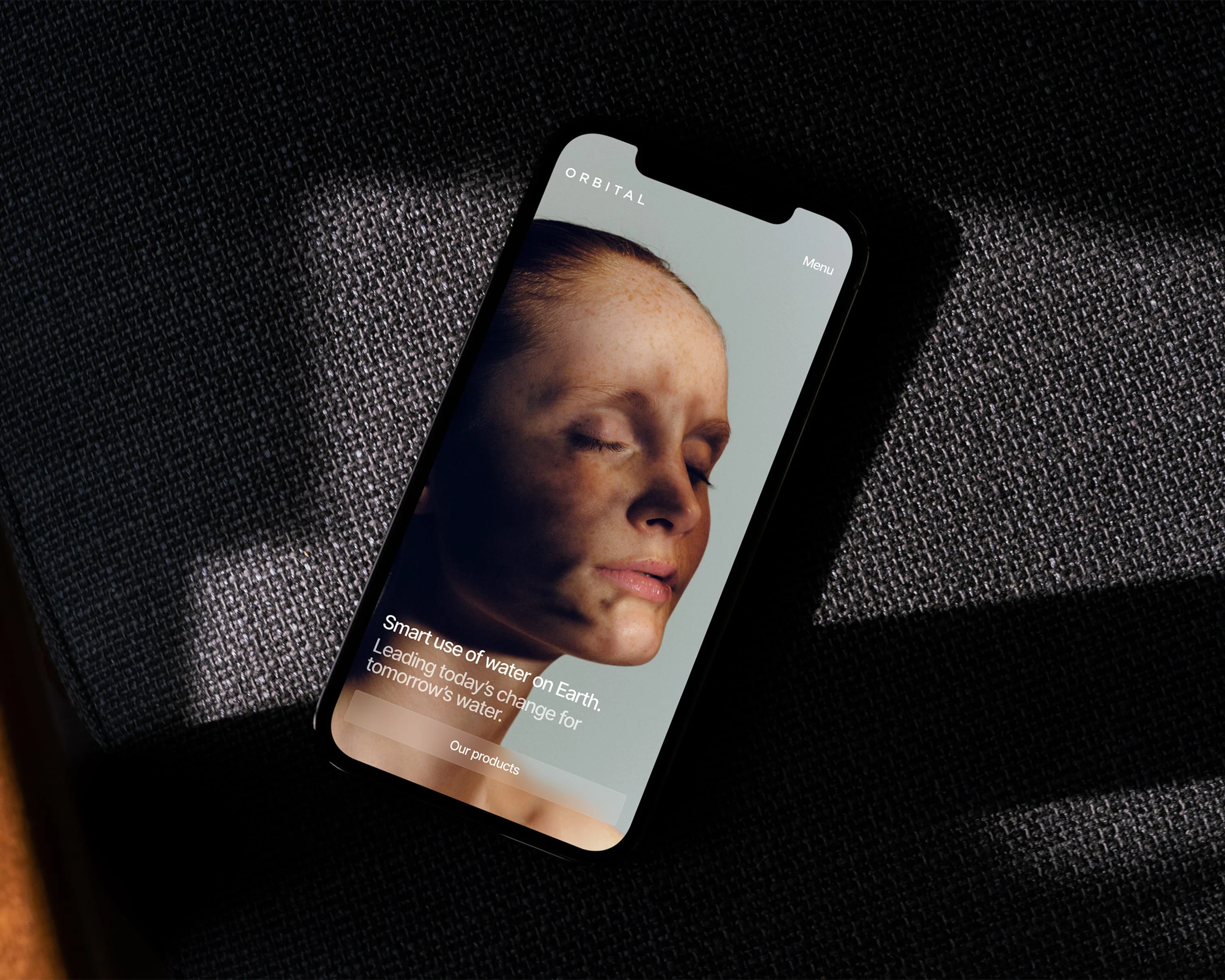 A smartphone displays the ORBITAL website featuring a person's dirt-streaked face and text about smart water use, resting on a dark textured surface.